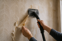 Person removing old wallpaper from a wall using a wallpaper steamer and scraper.
