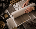 A top-down view of a person's hands unrolling a textured beige fabric or wallpaper sample over a styled collection of flooring and material swatches, including marble, wood, and metallic finishes.