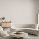 A modern living room scene featuring a curved, textured boucle sofa in a light beige/gray tone, a round marble coffee table with a gold bowl, a light wood sideboard with candles, and a textured wall.