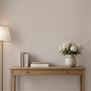 A styled interior scene showing a light wood console table against a warm beige wall, with a white lamp on the left and a ceramic vase of white flowers on the right. Books are stacked on the table.