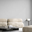 A modern living room scene featuring a beige tufted sofa, a round black coffee table with books, a small side table, and a matching ottoman against a minimalist light gray wall.
