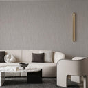 A modern living room featuring a textured light gray wall with a vertical gold LED light strip, a cream-colored sectional sofa with dark accent pillows, a marble-top coffee table, and a curved cream armchair.