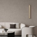 A modern minimalist living room featuring a curved white sofa with neutral throw pillows, a marble-top coffee table, and a sleek vertical gold wall sconce mounted on a textured warm gray plaster wall.